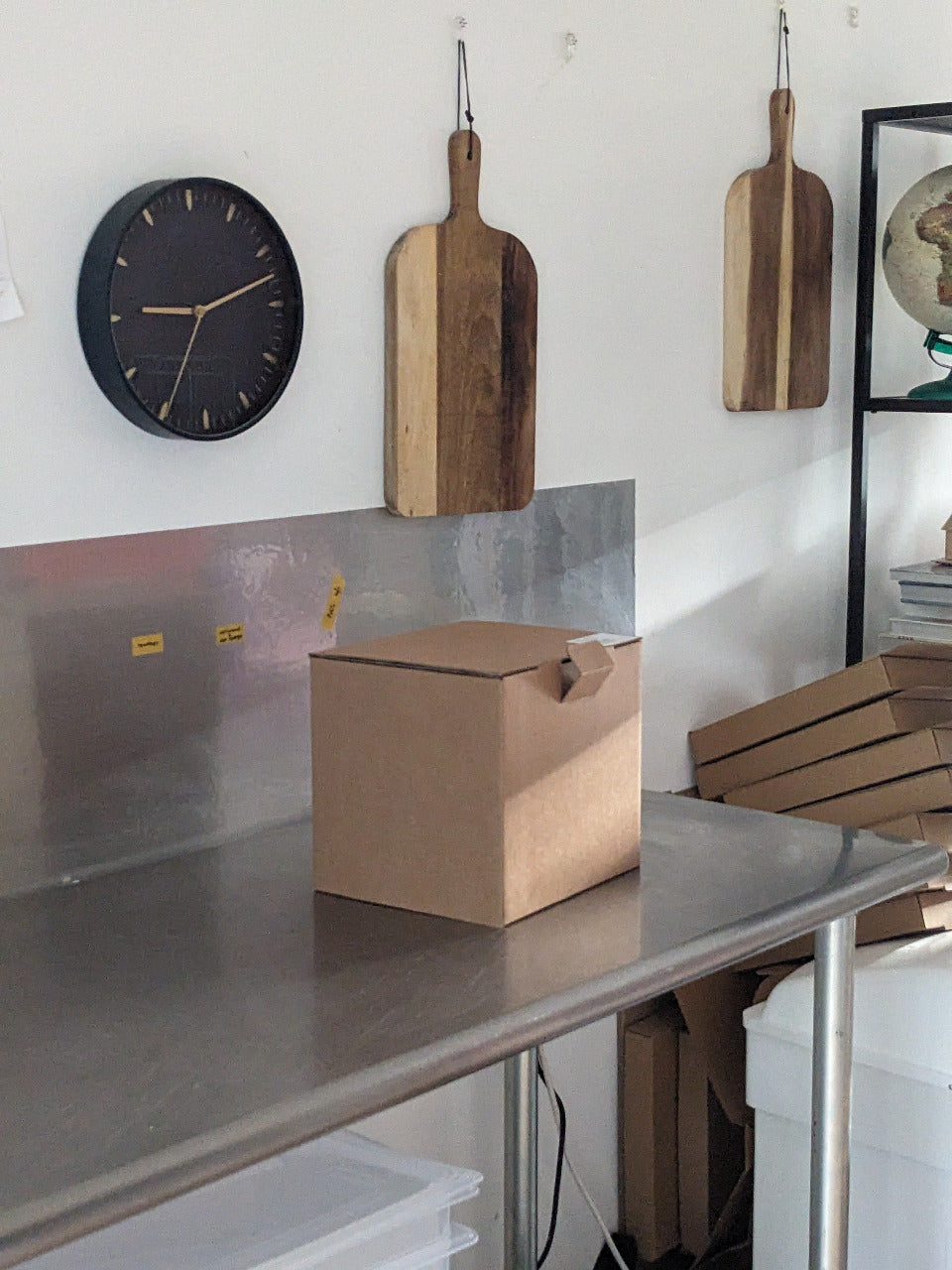 Cardboard box on a metallic surface with wooden cutting boards and a clock in the background.