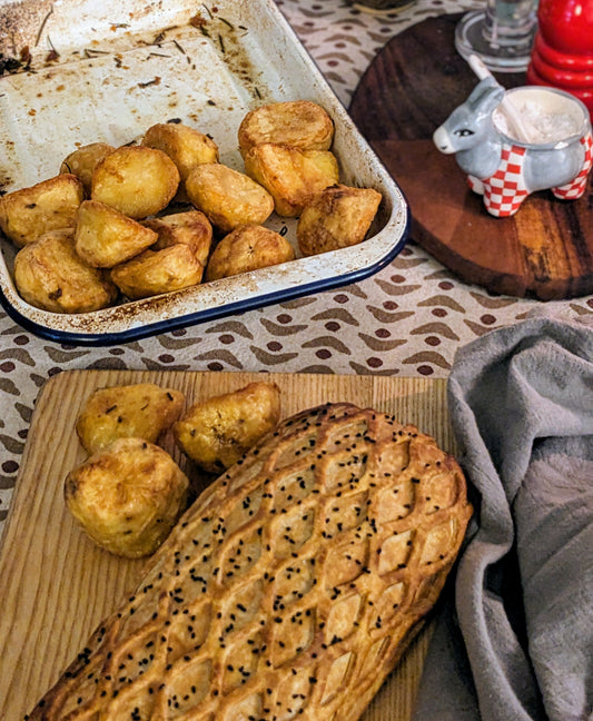 Uncut vegan mushroom and squash wellington with roast potatoes and sides on a patterned festive tablecloth.
